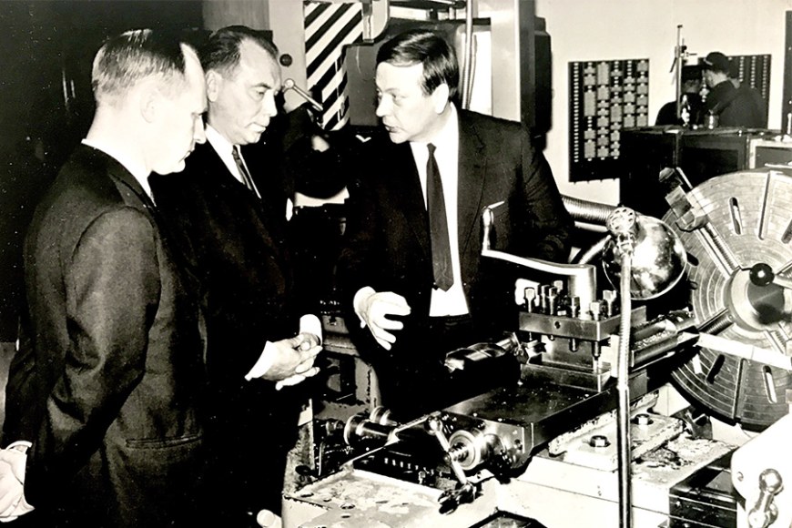 Mustavalkoisessa valokuvassa kolme miestä tarkastelee venttiilien valmistukseen käytettäviä laitteita tehdastilassa.