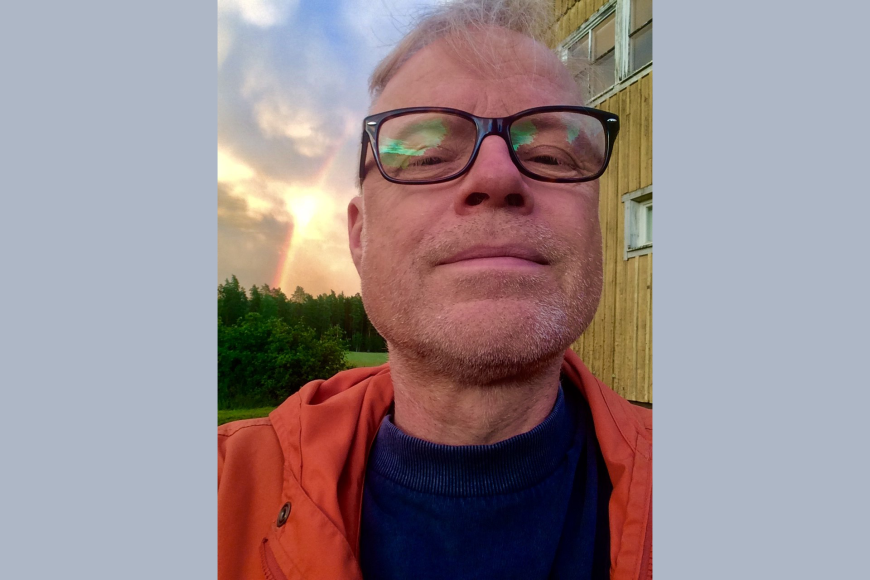 Professori Esa Kirkkopelto, selfie ulkona, taustalla maalaismaisemaa.