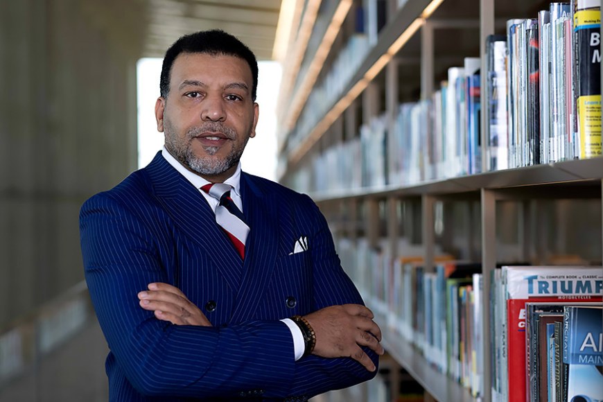 Mohamed Boufarss is standing next to a long bookshelf with his arms crossed. He is wearing a navy blue pinstripe jacket.