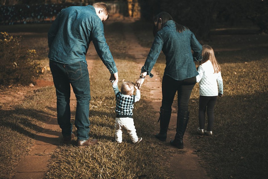 Neljä henkilöä kävelee eteenpäin. Vanhemmat pitävät pientä lasta käsistä ja toinen lapsi seuraa vierellä. 