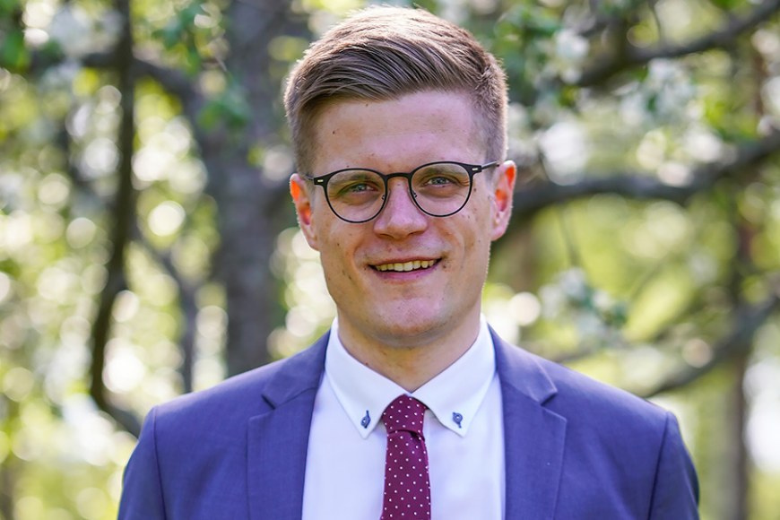 Rintakuva Rikhard Mäki-Heikkilästä, jolla on yllään sininen pikkutakki, valkoinen kauluspaita ja viininpunainen kravatti. Taustalla on lehtipuita.