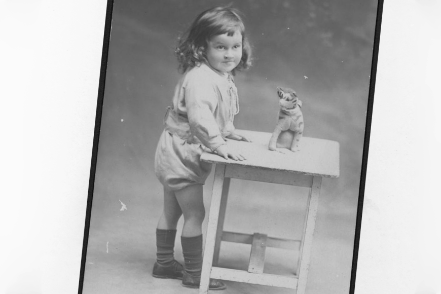 Vanha valokuva, jossa pieni lapsi seisoo pöydän ääressä ja pöydän päällä on lelutiikeri.