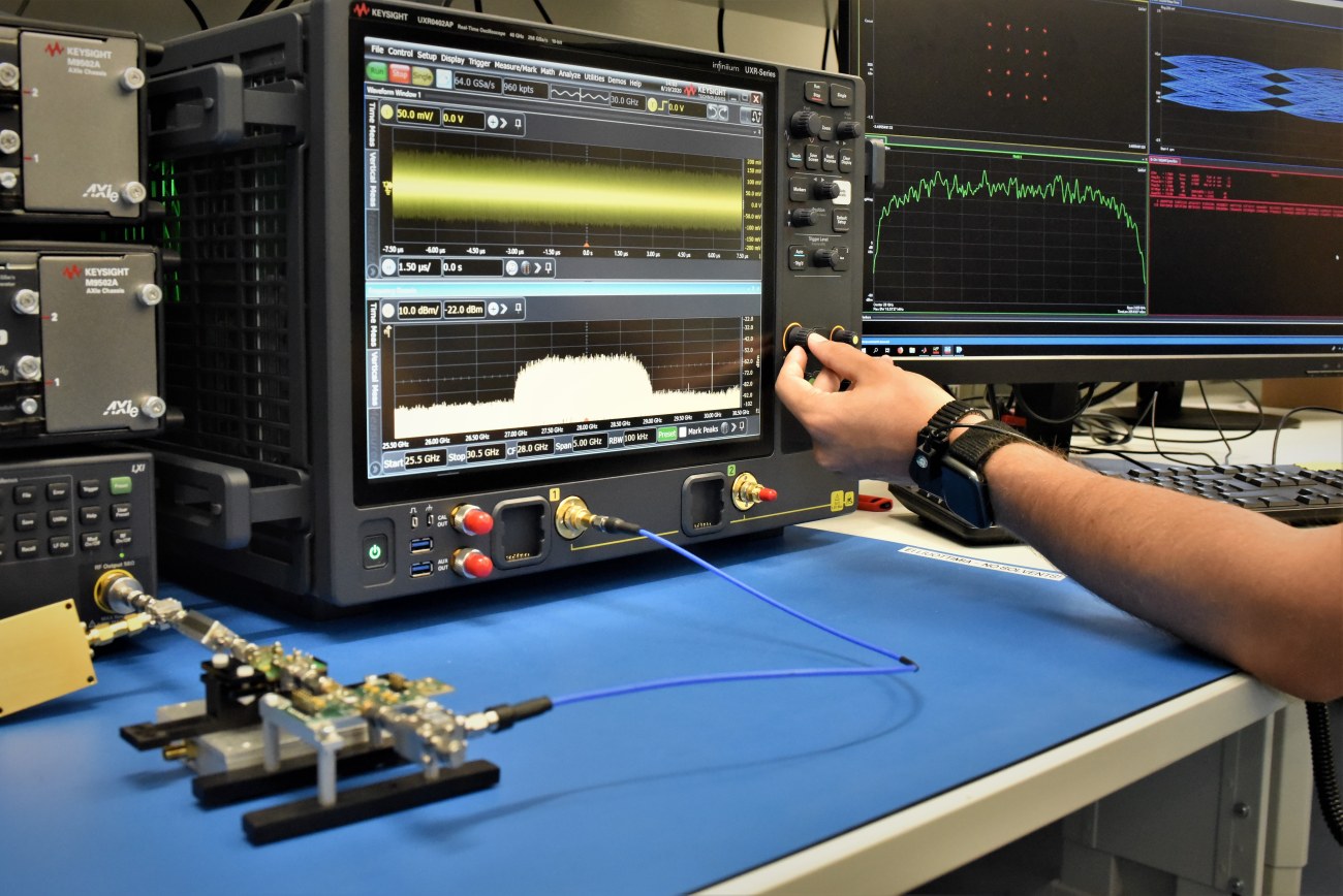 Measurement of a test RF board with Keysight real-time oscilloscope showing a center frequency of 28 GHz. A hand is adjusting one knob to fine tune the visualization.