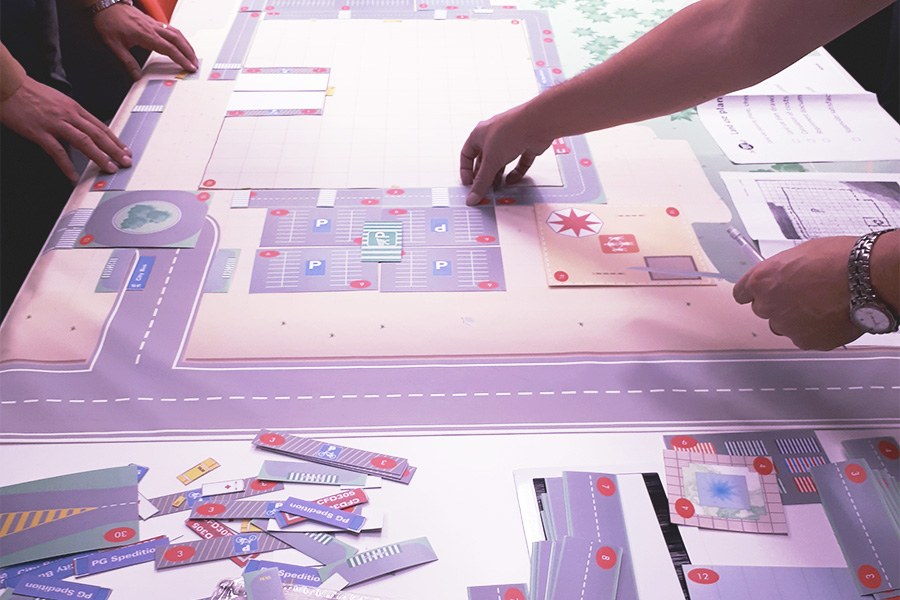 Pöydän päällä oleva simulaatiopelilauta, jonka ympärillä pelaajien käsissä pelikortteja.