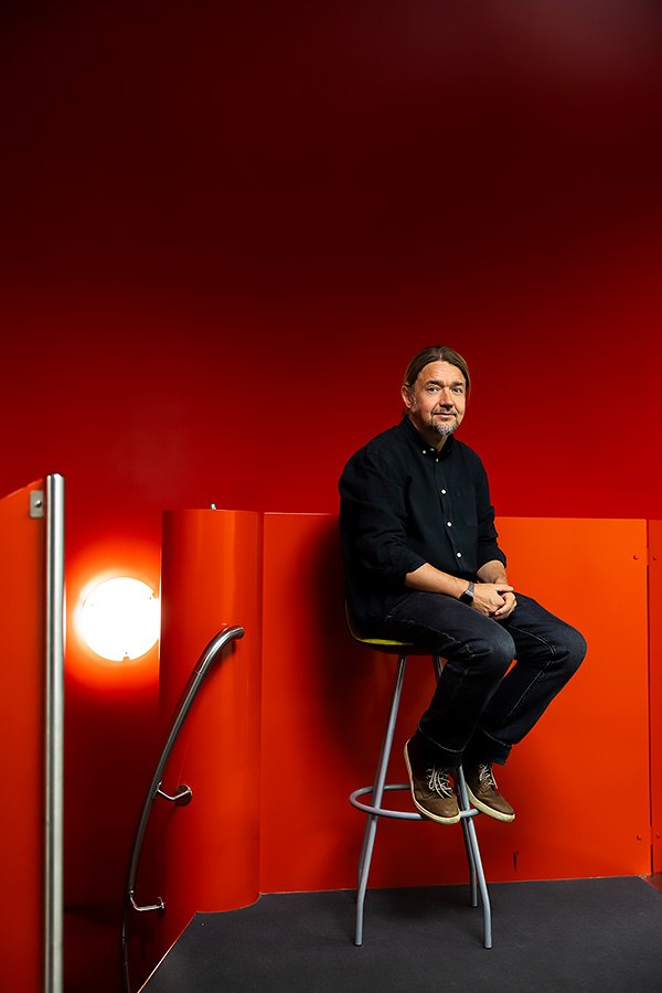 Markku-Juhani Saarinen sits on a high stool with his hands in his lap. The surrounding room is red.
