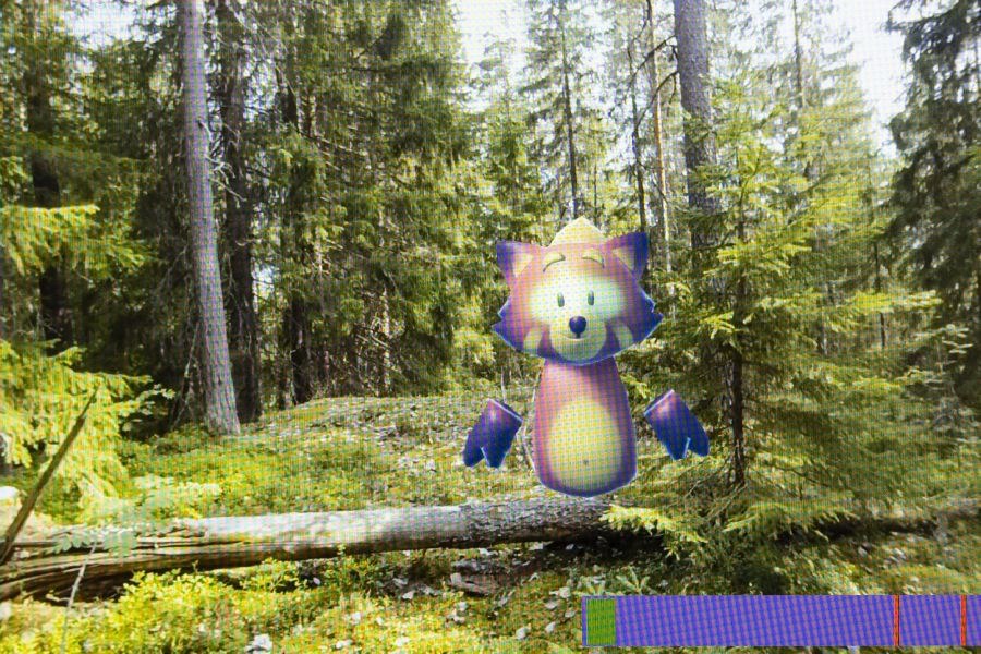 VR-sovelluksen näkymänä suomalainen metsämaisema. Taka-alalla vihreitä kuusipuita. Kuvan poikki on kaatunut puu sammalaiselle maalle. Kaatuneen puun päällä on digitaalinen kettuhahmo.