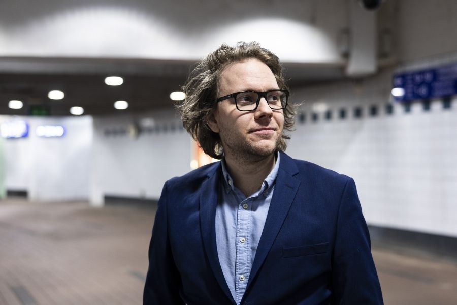 Esa Juntunen katsoo kamerasta oikealle yläviistoon. Hän seisoo metrokäytävällä ja hänellä on yllään tummansininen pikkutakki ja vaaleansininen kauluspaita. Esalla on mustapokaiset silmälasit ja laineikkaat ruskeat hiukset.