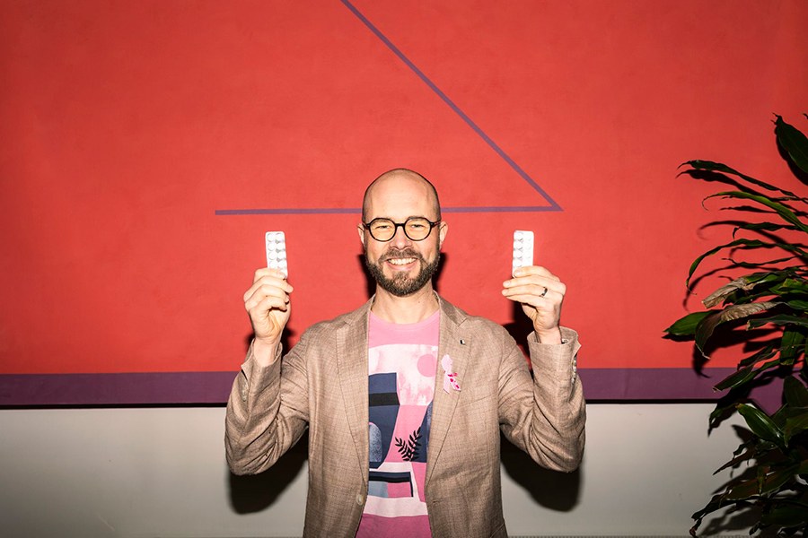 A person stands smiling in front of a red wall, holding up medical strips in they hands.