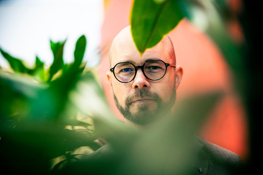 A person with glasses stands behind the leaves of a green plant and looks at the camera.