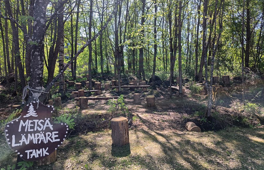 Metsäkaistale, jossa kyltti Metsälämpäre ja puupöllejä istuskeluun. 