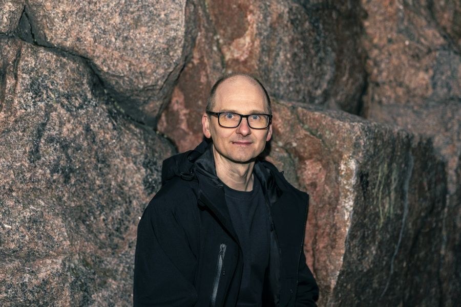 Silmälasipäinen henkilö seisoo kallion edessä mustassa takissa ja puserossa.