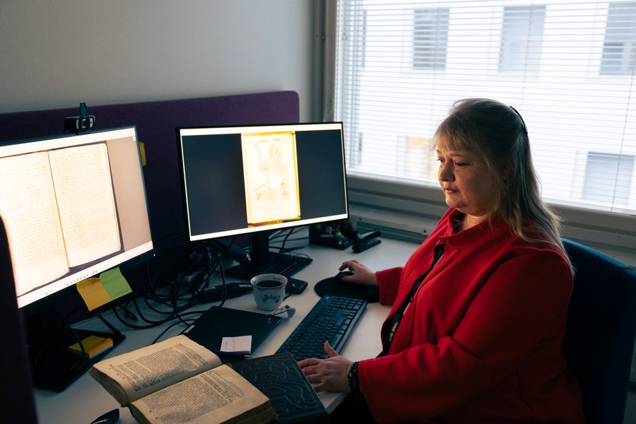 Raisa Toivo työpöytänsä äärellä. Näytöiltä näkyy vanhoja tekstejä ja pöydällä vanha kirja avoinna.