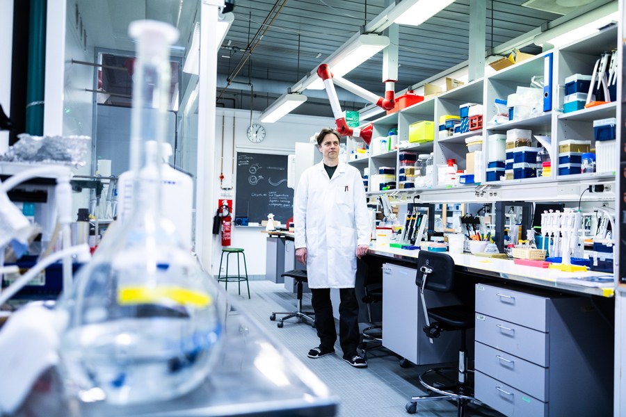 Valkotakkinen mies seisoo laboratoriossa, kuvan etualalla testimalja.