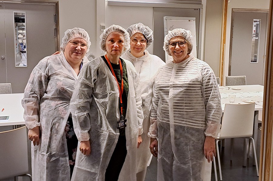 Neljä naista keittiötiloissa pukeutuneina suojahattuihin ja -kaapuihin. 