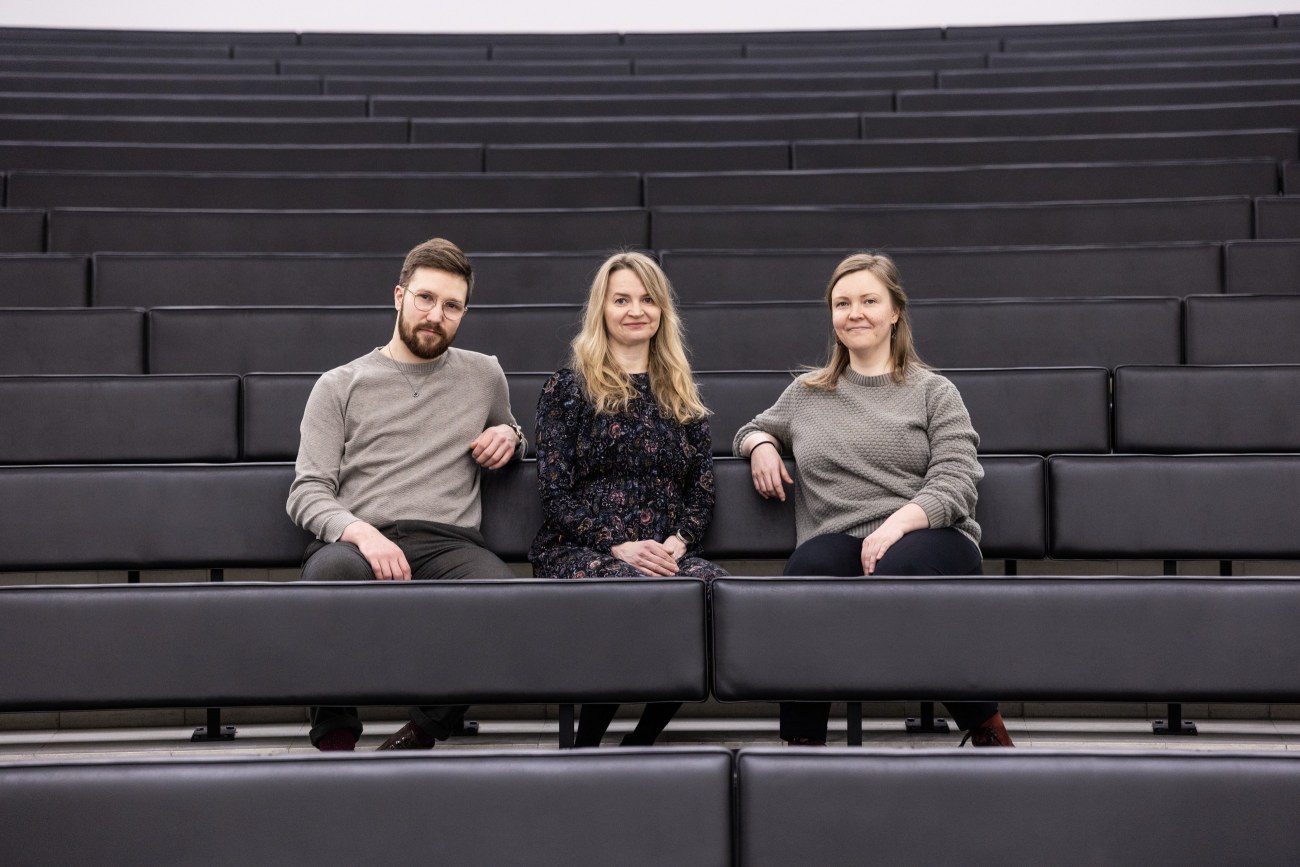Kolme henkilöä istuu juhlasalin penkeillä ja katsoo kameraan.
