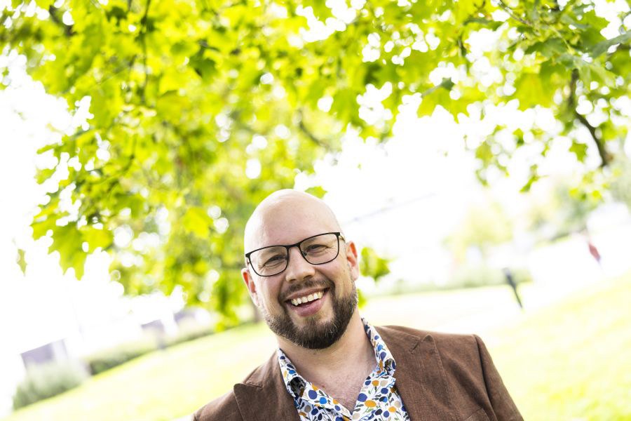 Hymyilevä henkilö ruskeassa puvuntakissa, taustalla näkyy puun lehtiä.