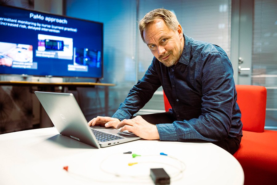 A person sits at a laptop computer and looks at the camera with a smile.