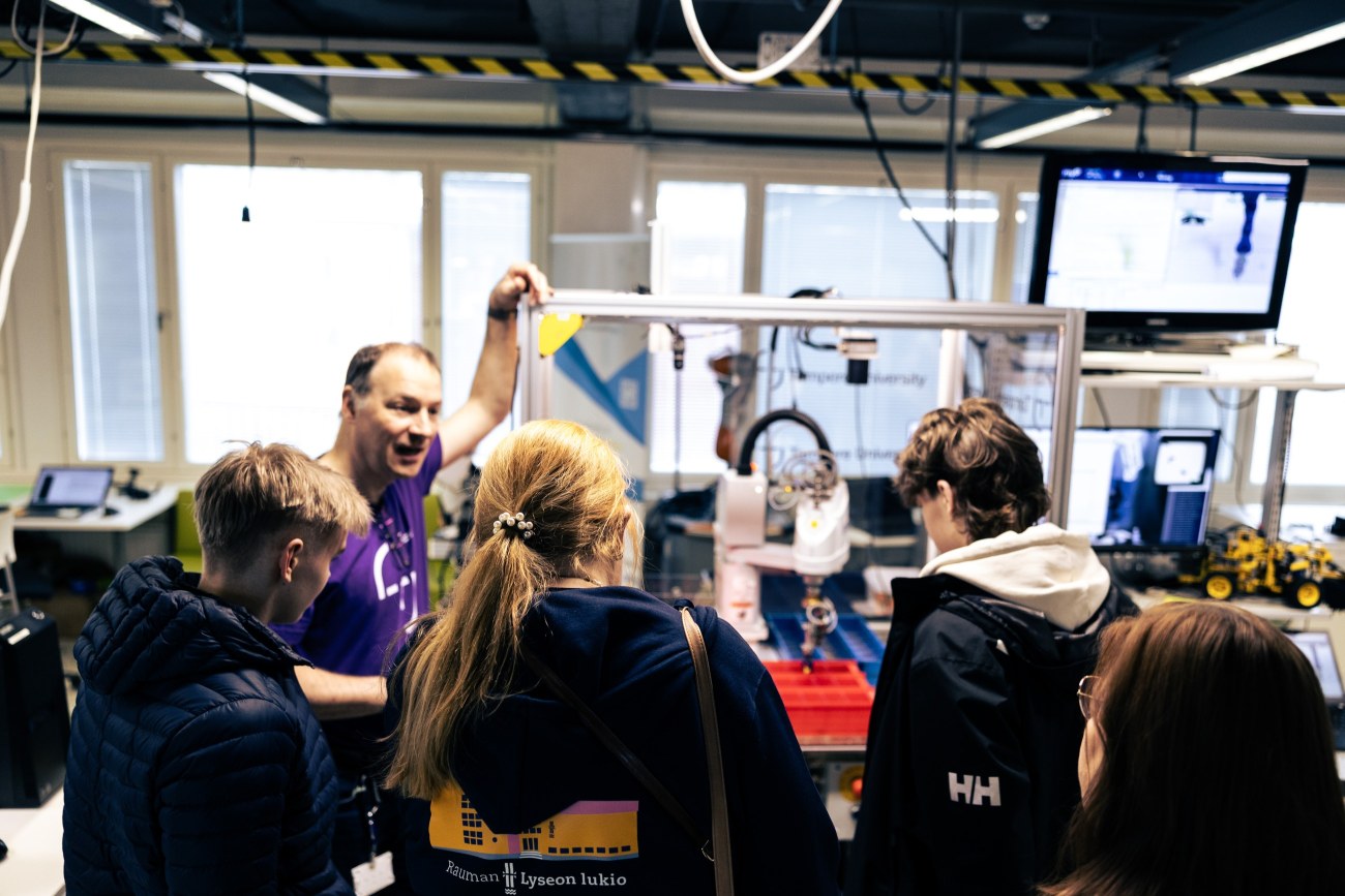 Neljä opiskelijaa selin kameraan, opettaja keskustelee heidän kanssaan samalla nojaten laatikkoon, jonka sisällä on robotti.