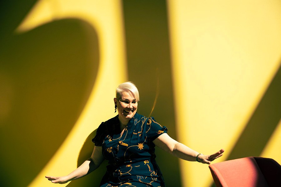 Tummansiniseen kuvioituun mekkoon pukeutunut henkilö heilauttaa käsiään kuin tanssissa. Taustalla keltainen seinä, johon lankeaa voimakkaita varjoja.