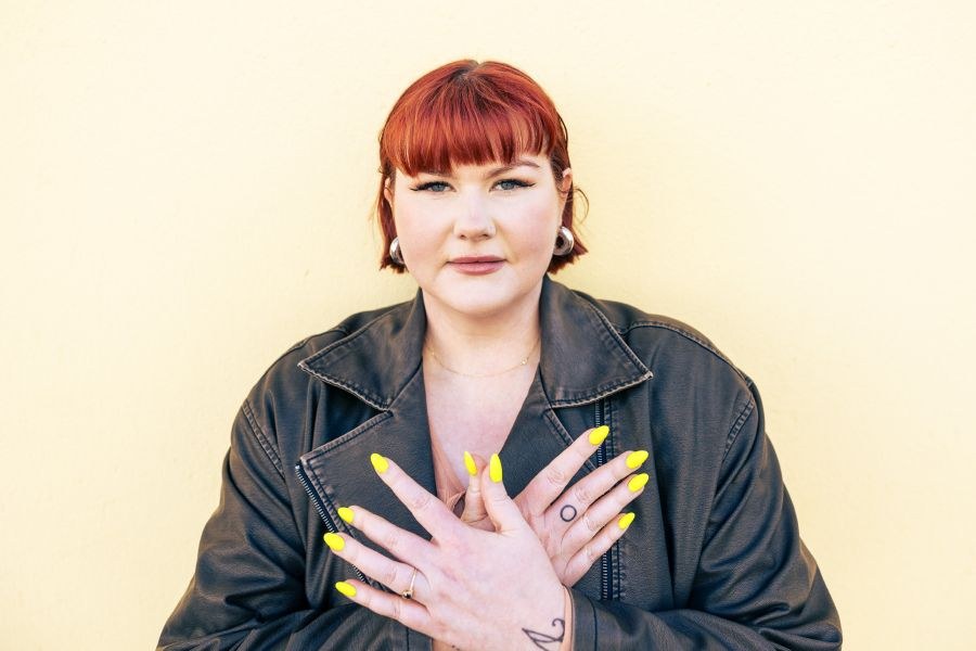 Punatukkainen nainen seisoo keltaisen seinän edessä ja katsoo kameraan. Hänellä on musta nahkatakki ja kädet, joissa on kirkkaankeltaiset kynnet, ovat päällekkäin, kämmenselkä kameraa kohti.