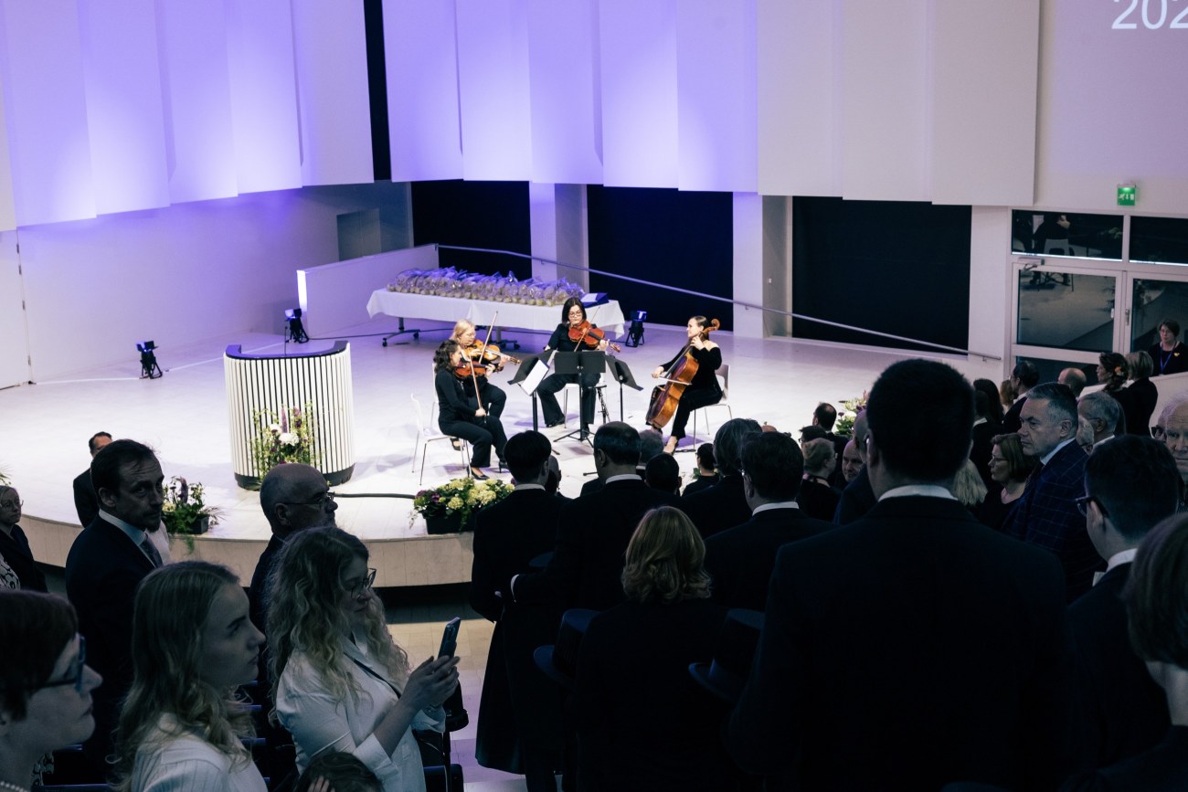 Juhlasalin lavalla neljä jousisoittimen soittajaa, kuvan alareunasta näkyy kuinka professorit kulkevat kulkueena kohti lavaa.