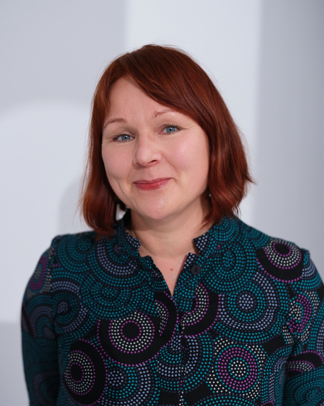 Anne Leppäjärvi hymyilee puolilähikuvassa, katse kameraan, taustalla valkoinen seinä.