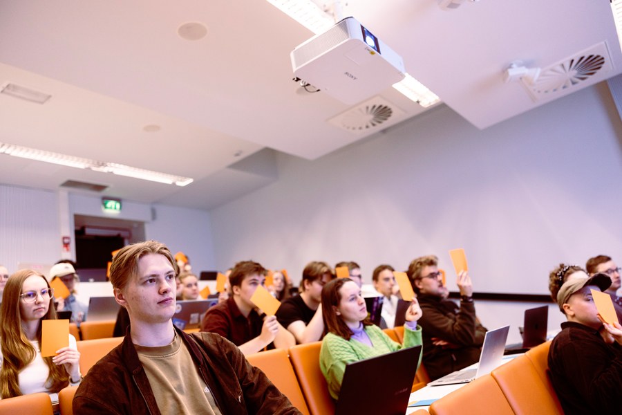 Opiskelijat kuuntelevat luentoa. Käsissä näkyy nostettuja äänestyslappuja.