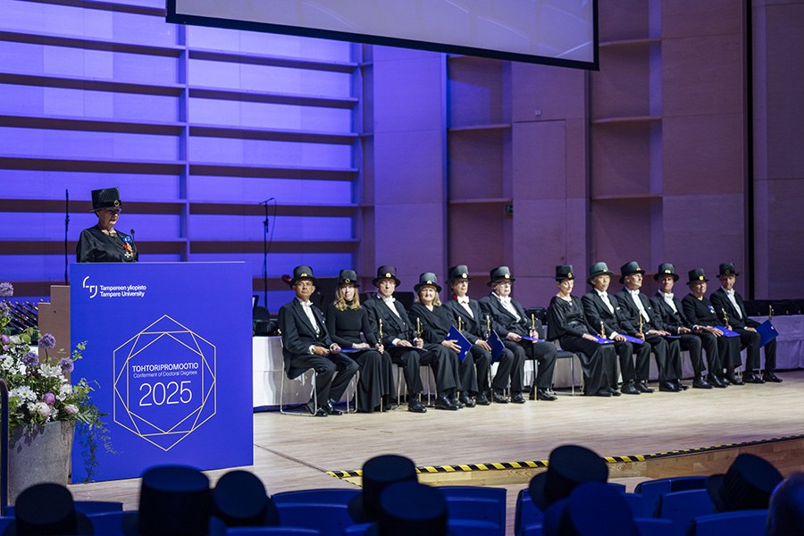 Kunniatohtorien edustaja pitää puhetta ja muut kunniatohtorit istuvat esiintymislavalla