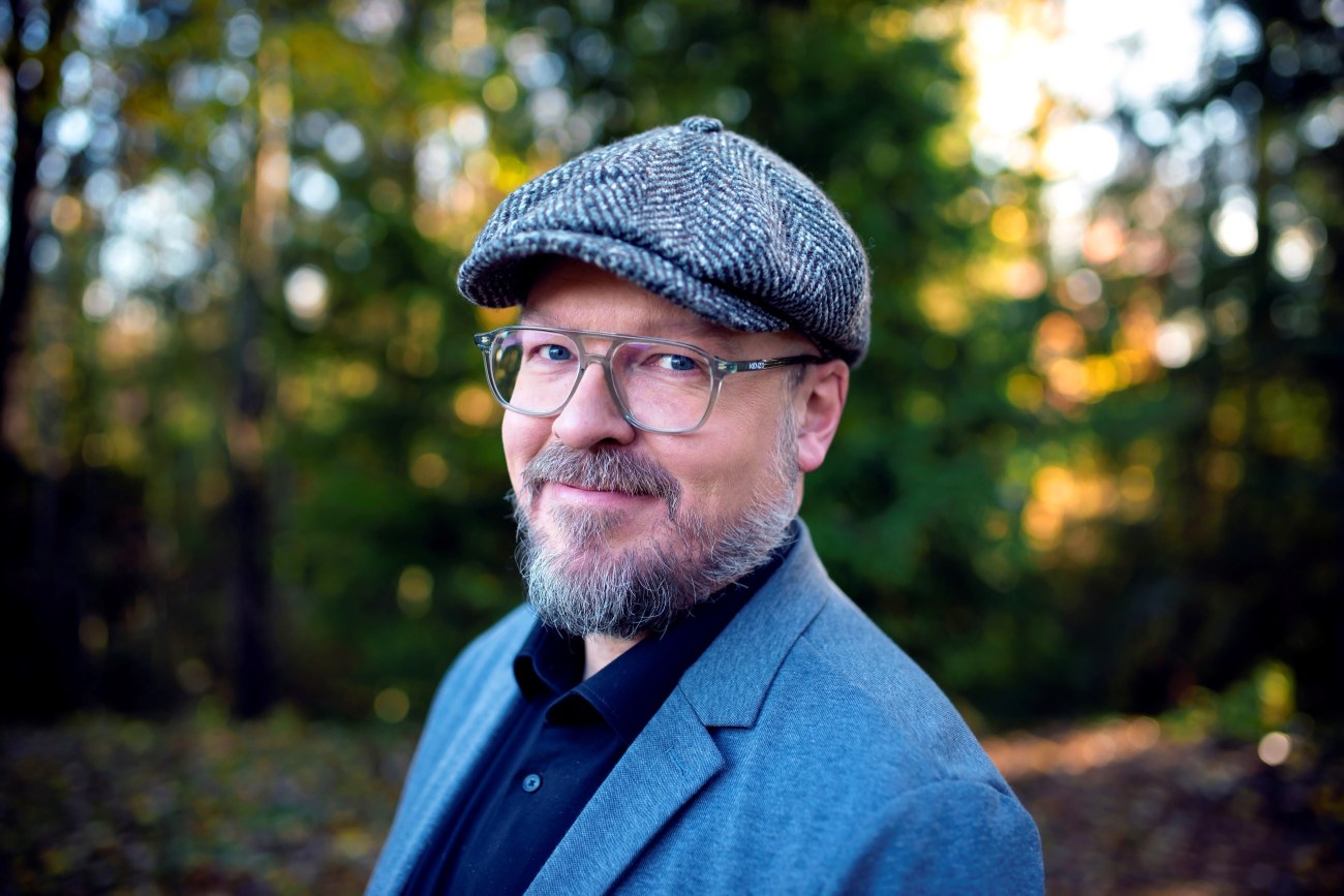 Mikko Hautakangas hymyilee lähikuvassa, katsoo kameraan, taustalla puita ja auringonvaloa.