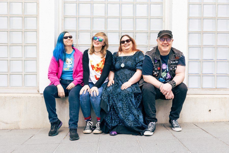 Neljä hymyilevää pelitutkijaa istuu katsoen kameraan, taustalla valkoinen lasiseinä.