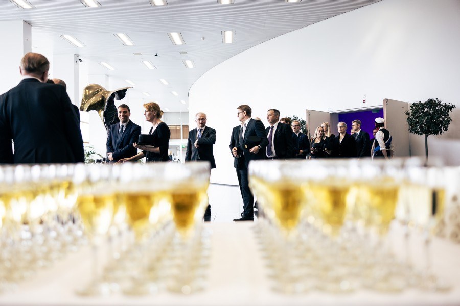 Etualalla kuohuviinilaseja pöydällä. Takana juhlasalista pois tulevia avajaisvieraita.