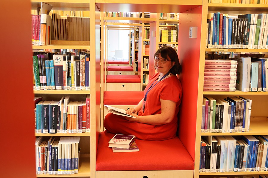 Nainen istuu kirjastossa ja pitää kirjaa sylissään. 