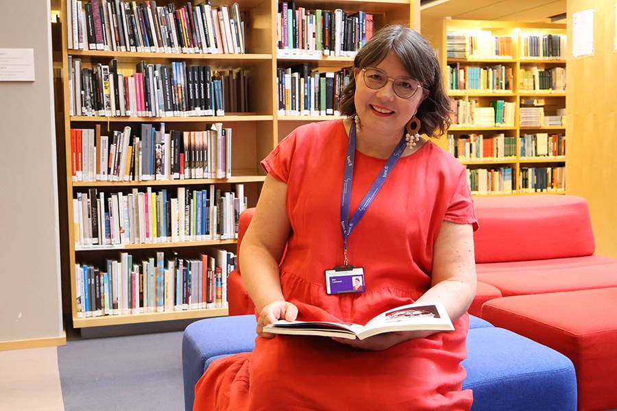 Nainen istuu kirjaston penkillä ja pitelee kirjaa sylissään. 