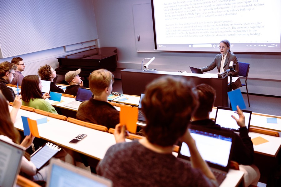 Students in a lecture hall holding blue and orange pieces of paper in their hands. The teacher watches from the front.