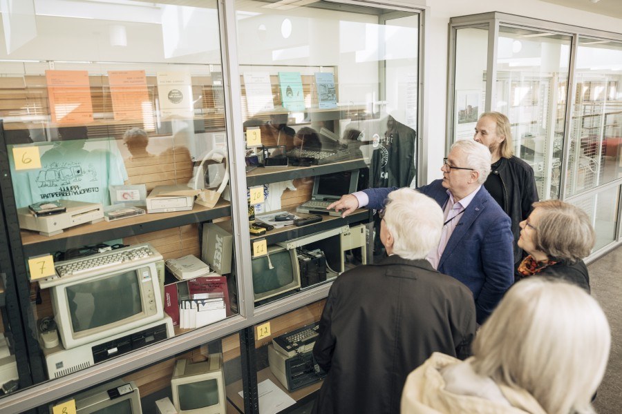 Tietotekniikan museonäyttelyvitriinissä vanhoja laitteita. Etualalla juhlavieraita katsomassa vitriiniin.