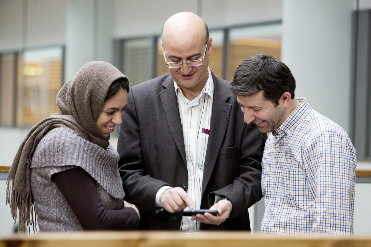 Kolme henkilöä pöydän takana tutkimassa keskimmäisen kädessä olevaa älypuhelinta.