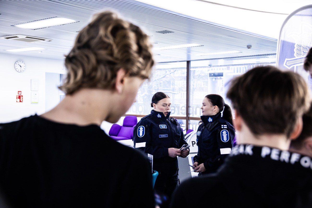Etualalla kaksi henkilöä selkä kuvaajaan päin. He katsovat kuvan keskellä olevaa kahta henkilöä, joilla on päällään poliisin univormu. 