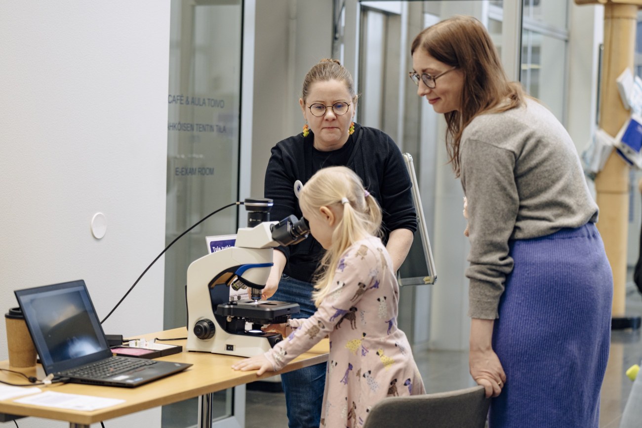 Lapsi kurkistaa mikroskooppiin. Kaksi aikuista henkilöä nojautuu kohti lasta ja hymyilee.
