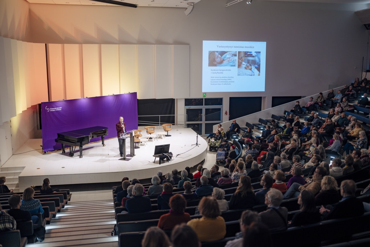 Juhlasalin istumapaikat ovat täynnä yleisöä. Lavalla on violetti taustakangas, jonka edessä seisoo henkilö. 