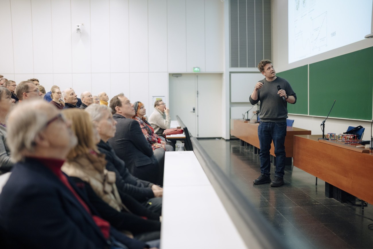 Yleisö istuu auditoriossa, ja katsoo liitutaulujen yläpuolella olevalle valkokankaalle heijastettua kuvaa. Heidän edessään seisoo henkilö, joka katsoo myös valkokankaalle.