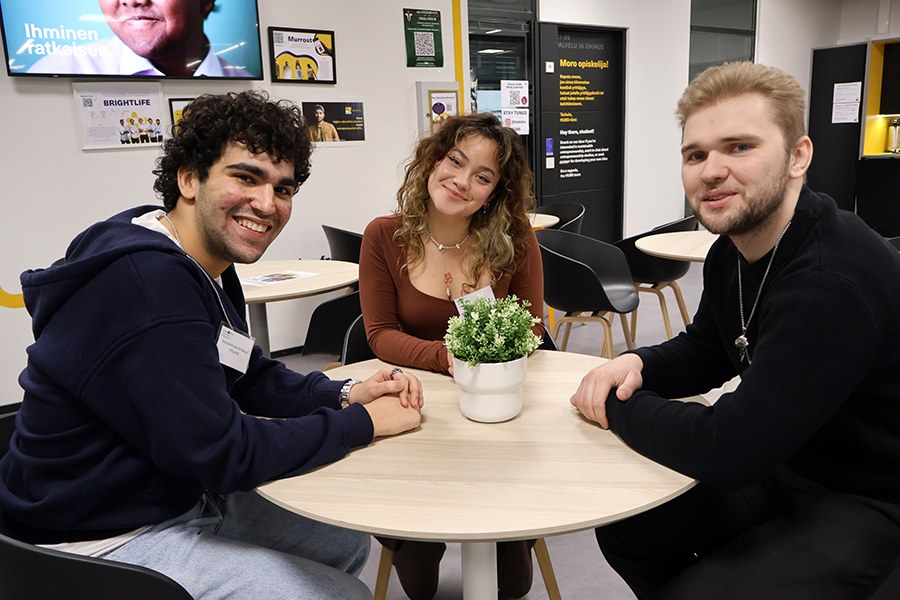 Kolme nuorta aikuista pöydän ääressä hymyilee ja katsoo kameraan. 