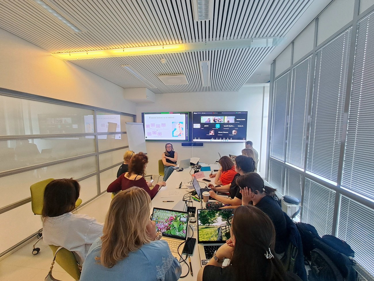 Students and staff sat around a table with 2 monitors showing slides and online students joining on zoom