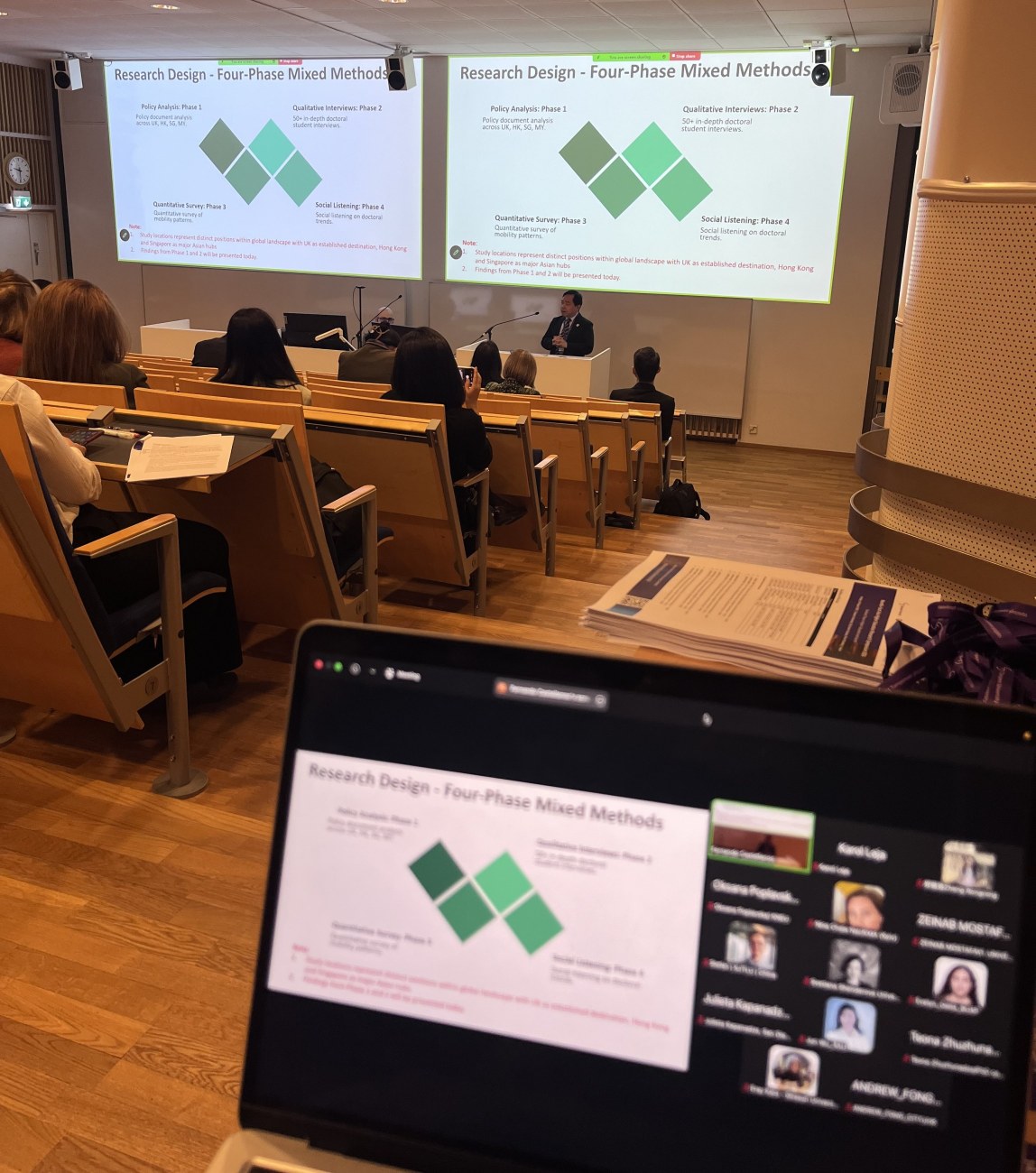 Conference room with a presentation ongoing and a view of a laptop screen in the foreground showing an ongoing Zoom stream