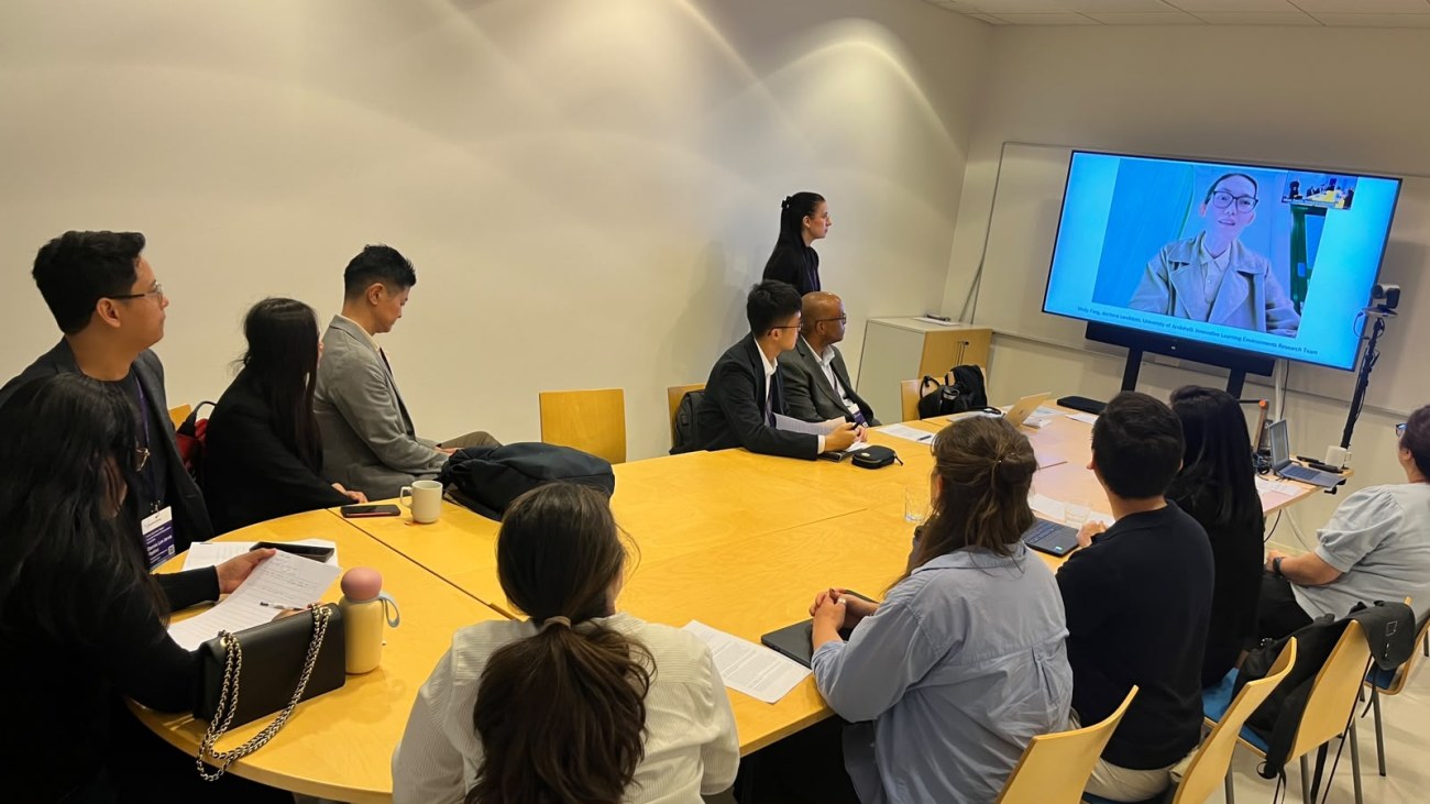 A parallel session ongoing with an online participant presenting on the screen in the back of the room and a group of academics sitting around a conference table