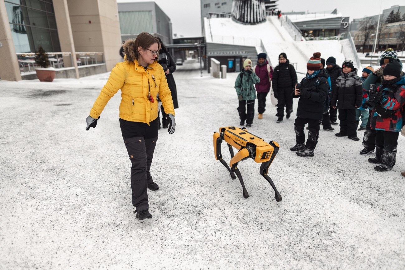 Henkilö keltaisessa talvitakissa kävelee keltaisen robottikoiran vierellä. Taustalla joukko lapsia seuraa.