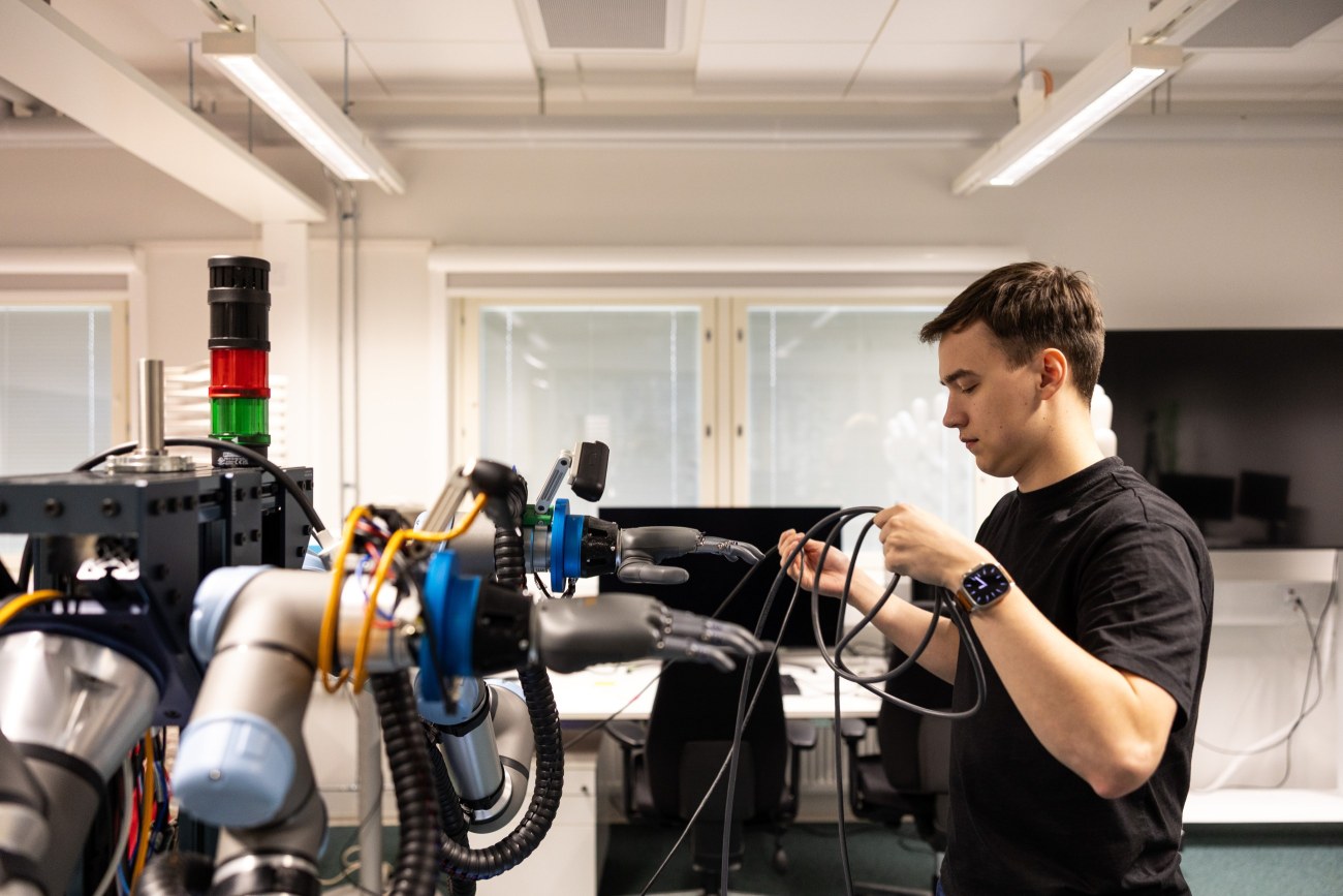 Ihminen seisoo robottikäsien edessä ja pitelee johtoja käsissään.