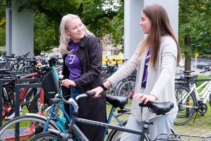 There are several bicycles beside the main building of Tampere university. Two female students are standing and holding their bikes while chatting with each other.