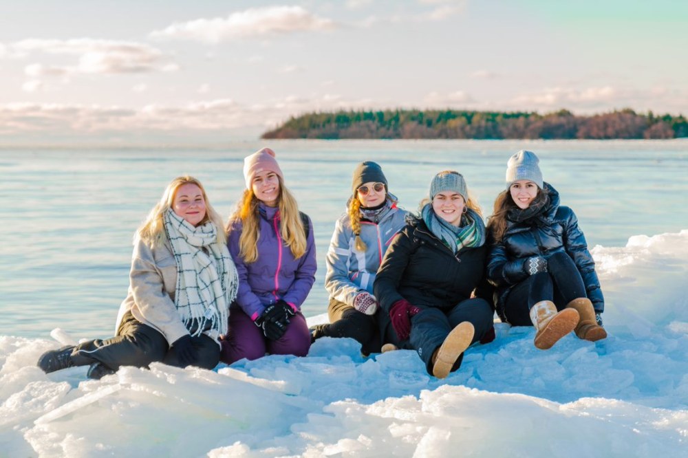 Meren rannalla talvisessa aurinkoisessa säässä jäisellä rannalla istuu viisi hymyilevää vaihto-opiskelijaa.